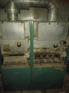 A pair of old, rusted industrial machines with green panels and silver ductwork await furnace repair in a dimly lit room.